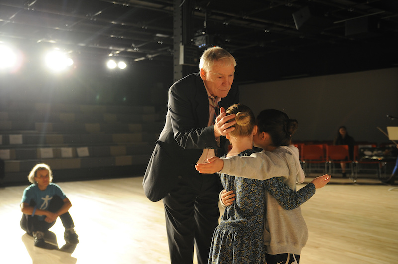 Two young girls embrace on the NDI stage, Jacques and another teacher coach them